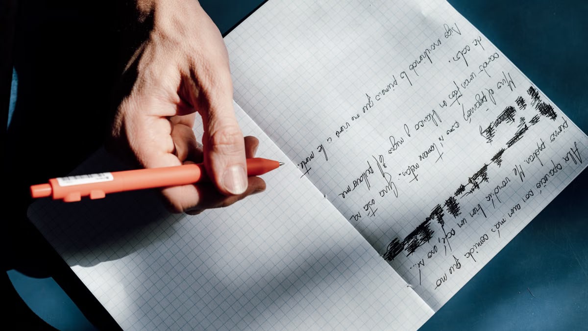 A person practicing handwriting using a red pen in a notebook during a sunny day in Buenos Aires.